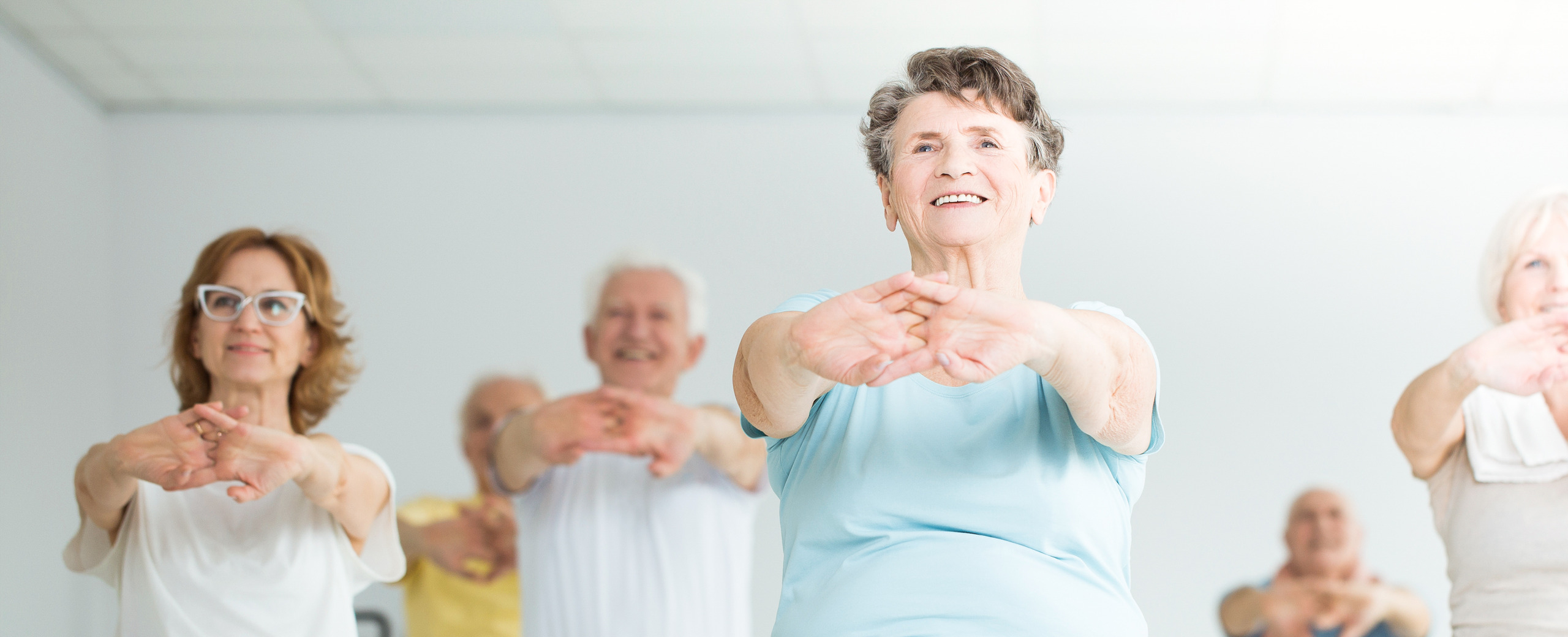 A group of older adults smiling and stretching their arms forward during a fitness or exercise class in a bright, airy room.