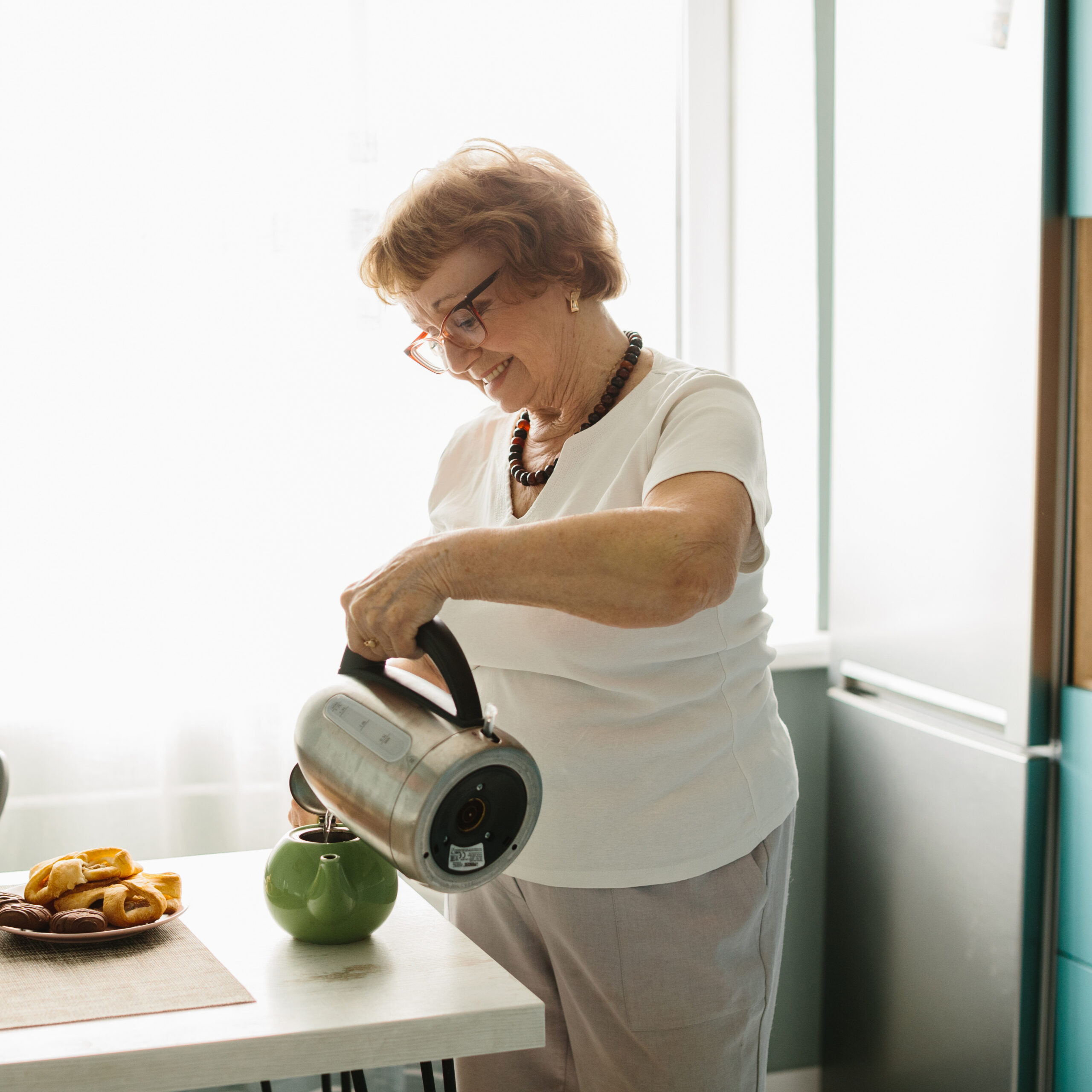 An elderly woman with glasses smiles as she pours hot water from an electric kettle into a green teapot on a kitchen counter with pastries on a plate nearby.