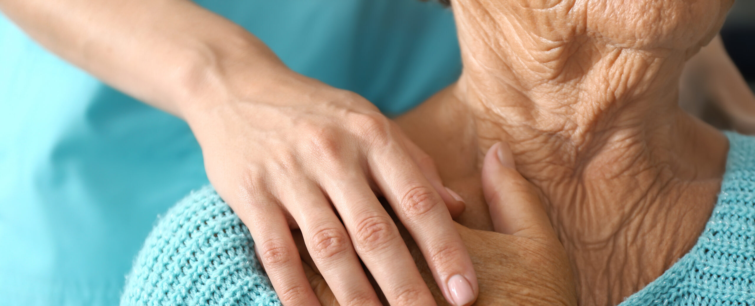 A younger person’s hand rests gently on the shoulder of an older adult wearing a knitted blue sweater and a wristwatch, conveying care and support.