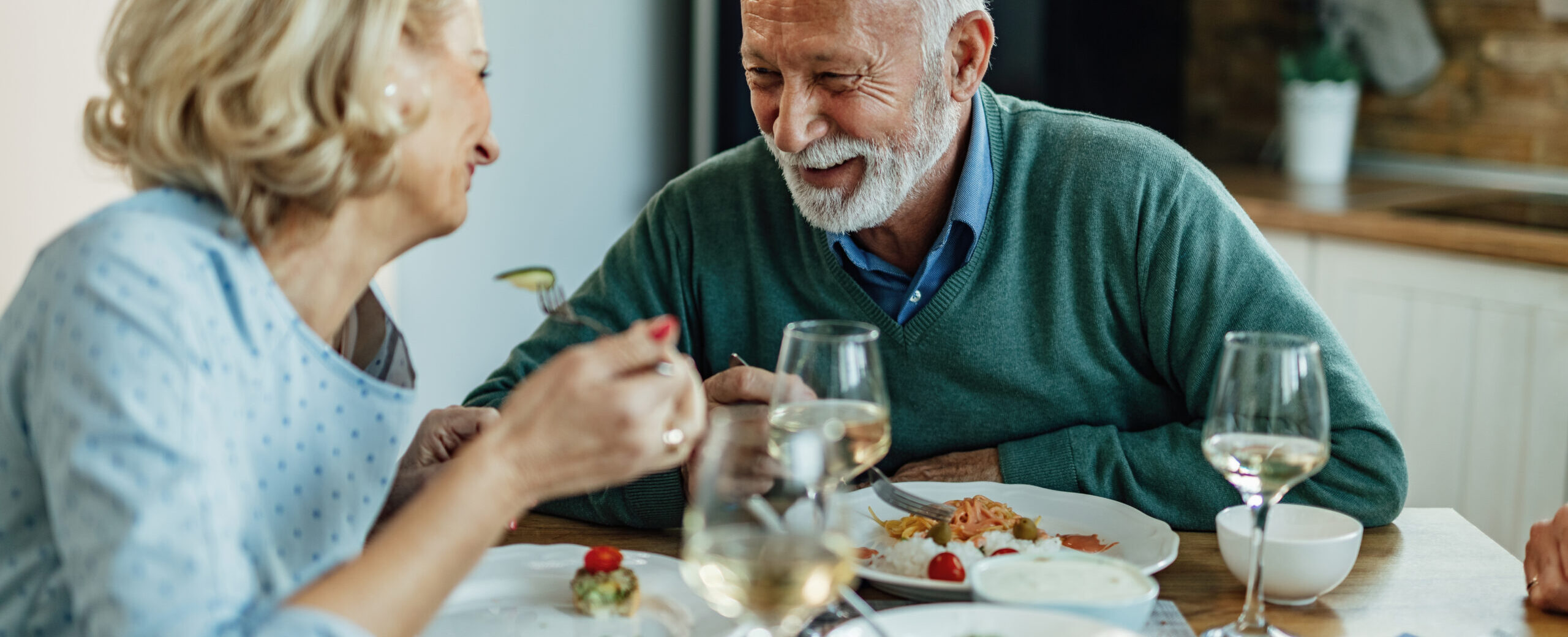 An older couple sits at a dining table, laughing and enjoying a meal together. There are plates of food and glasses of white wine on the table in a cozy, warmly-lit kitchen.