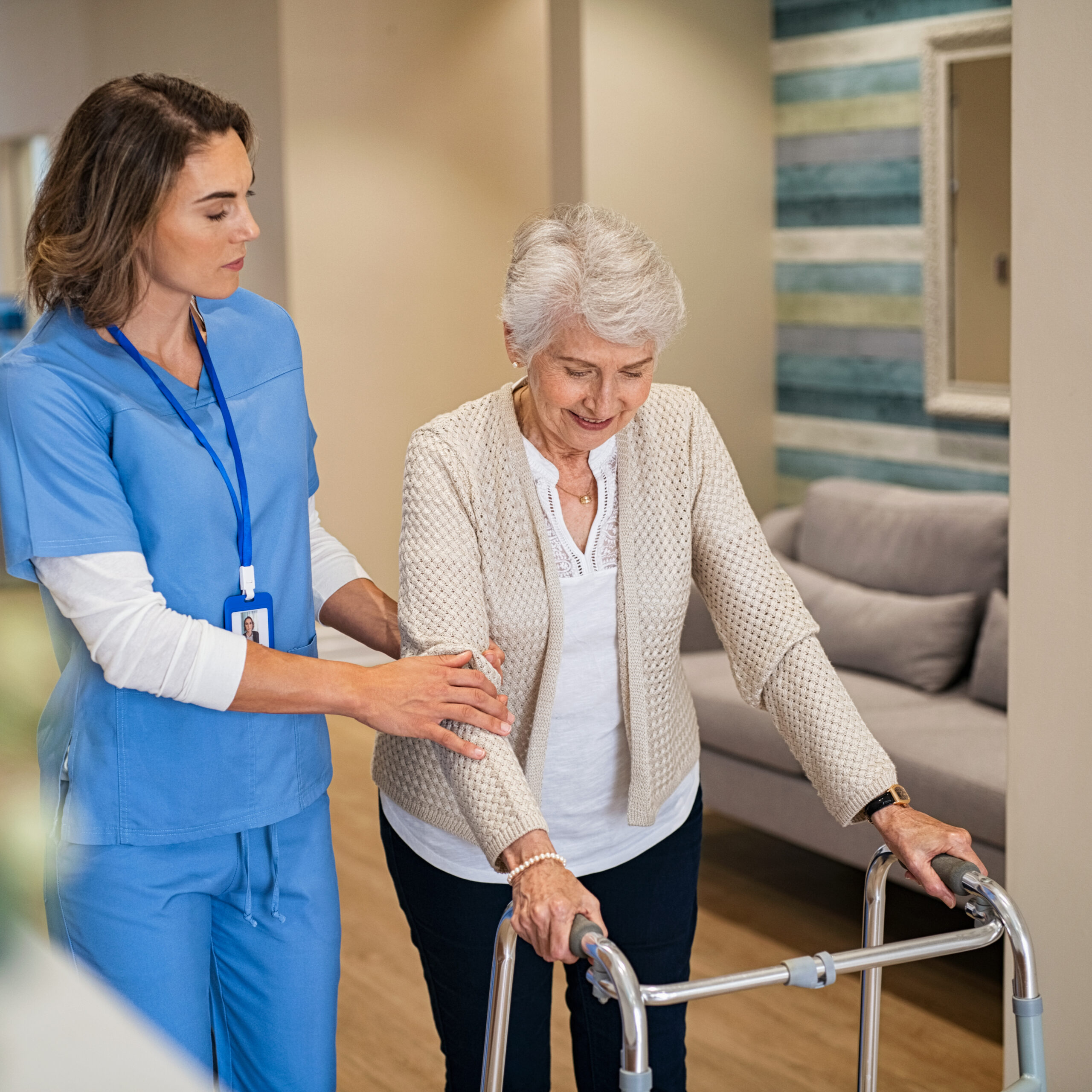 A nurse in blue scrubs assists an elderly woman using a walker in a well-lit, comfortable room with a couch and a striped wall. The nurse gently supports the woman, who is smiling during the activity.