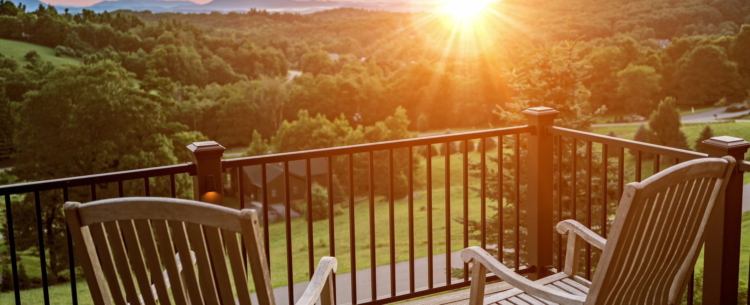 Two wooden chairs on a balcony overlook a lush, green landscape and rolling hills at sunset, with sunlight streaming over the mountains and trees in the distance.