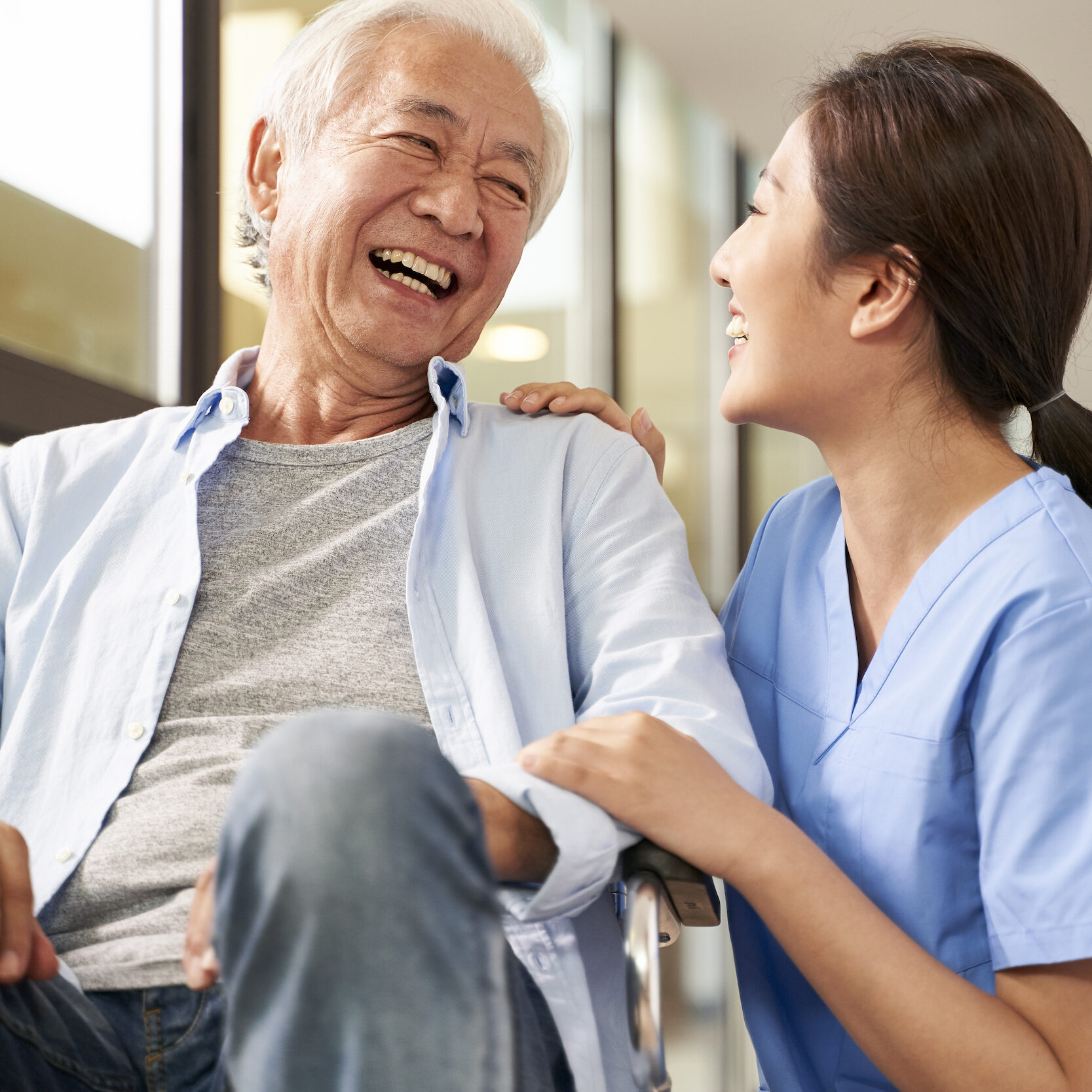 An elderly man in a wheelchair laughs joyfully while a young female caregiver in blue scrubs smiles and holds his arm, both sharing a happy moment in a bright, indoor setting.